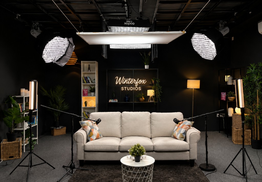 Three seater podcast studio with beige sofa, Winterfox Studios neon sign, overhead softboxes, and boom microphones at Winterfox Studios
